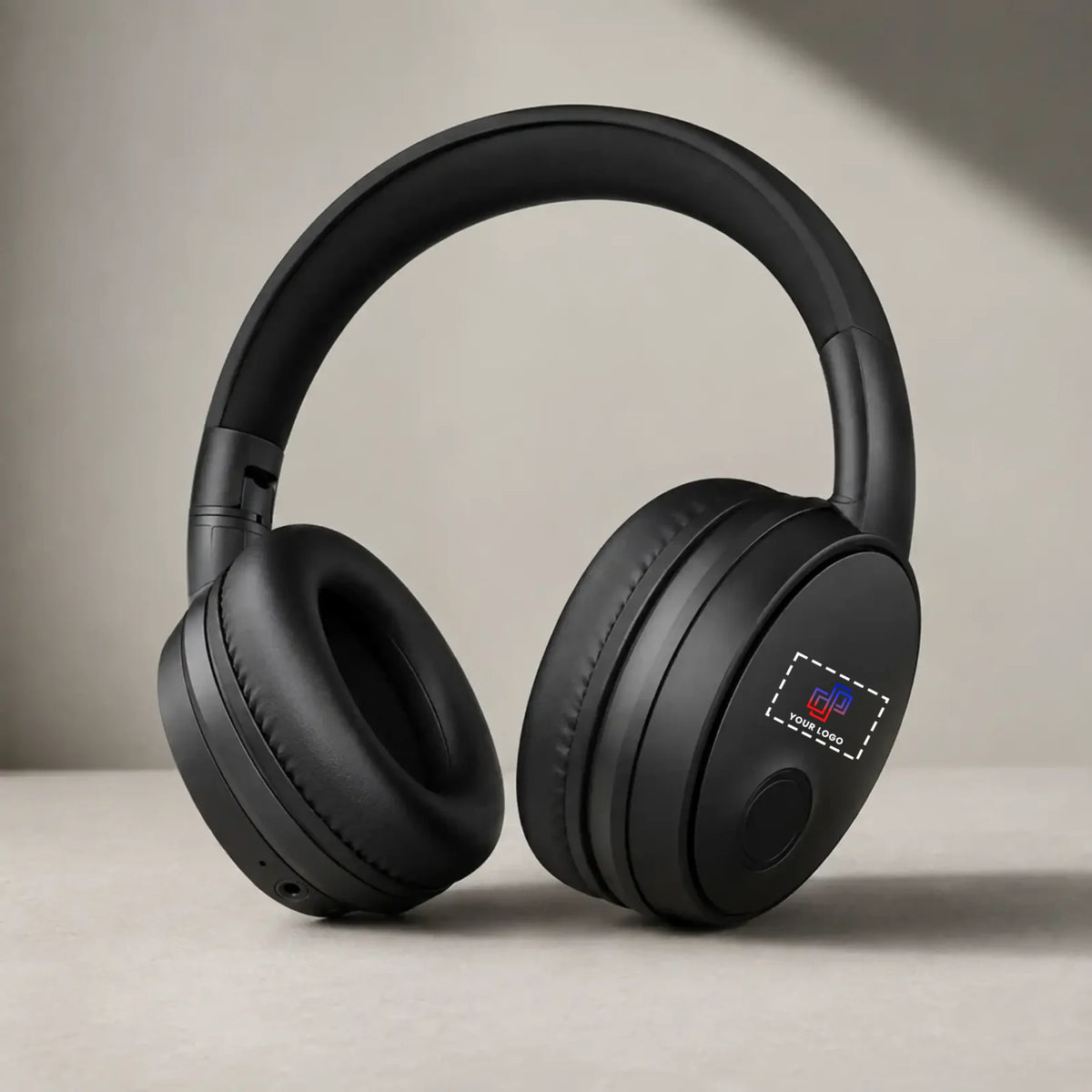 Black over-ear wireless headphones with cushioned ear cups and a padded headband, shown resting on a light beige surface against a neutral beige background.