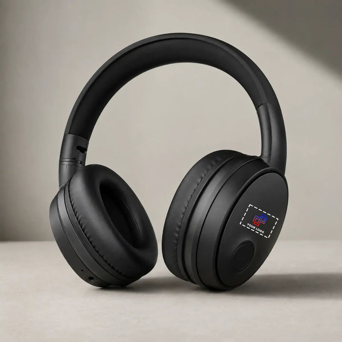 Black over-ear wireless headphones with cushioned ear cups and a padded headband, shown resting on a light beige surface against a neutral beige background.